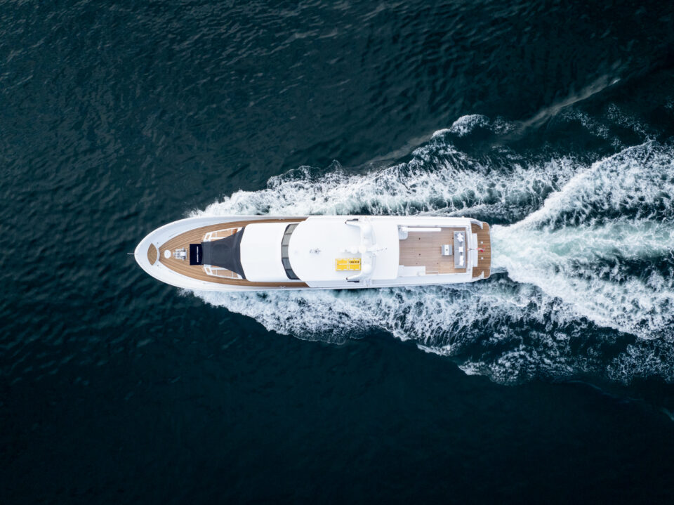 Aerial view of a white luxury superyacht cruising through dark blue water, creating waves and a foamy wake behind it. The yacht’s deck and sleek design are clearly visible—perfect for luxury superyacht hire or game fishing charters.