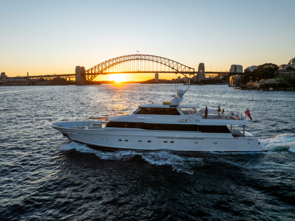 A large white yacht from Shore Thing yacht charter sails on the water at sunset, with the Sydney Harbour Bridge in the background and the sun setting behind it, casting a warm glow over the scene.