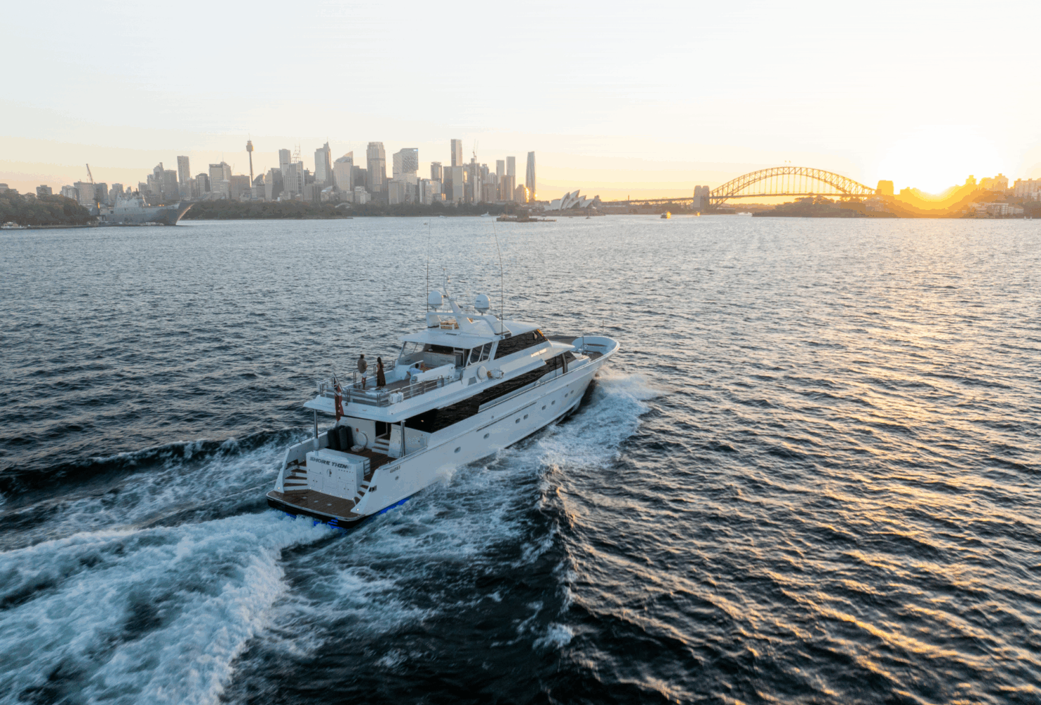 A large yacht from Shore Thing yacht charter sails through the water at sunset, with the Sydney city skyline and Harbour Bridge visible in the background.