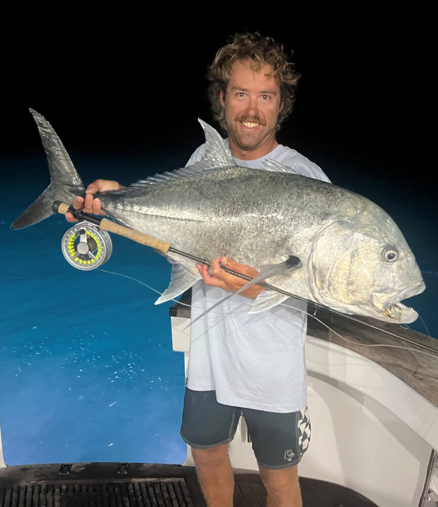 A man with curly hair, wearing a light shirt and shorts, smiles as he holds a large silver fish and fishing rod on a game fishing charter boat at night, with dark water in the background.