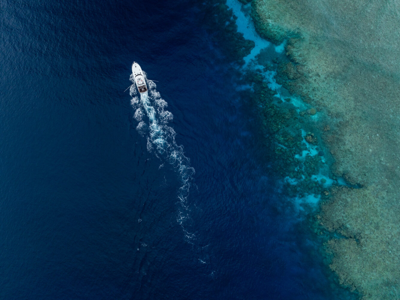 A white luxury superyacht travels through deep blue ocean water near a shallow, turquoise coral reef, leaving a visible wake behind. The scene is viewed from above.