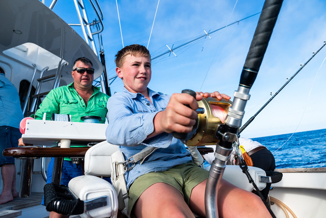 shore thing fishing charter drone and fishing images (2) A boy sits in a chair on a luxury superyacht, holding a fishing rod and reeling it in, while an older man in sunglasses watches behind him. The ocean and a clear blue sky are visible in the background.
