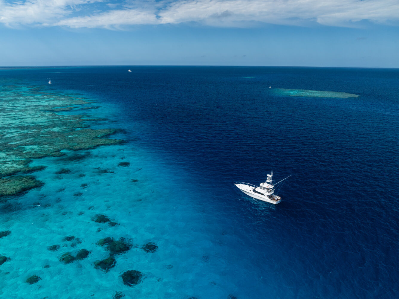 A luxury superyacht floats on clear blue ocean water near a coral reef, with small patches of reef visible below the surface and a few distant boats under a partly cloudy sky.