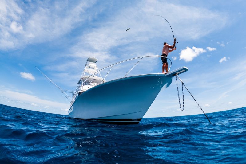 A person stands on the bow of a luxury yacht hire, holding a bent fishing rod over deep blue ocean water under a bright, partly cloudy sky.