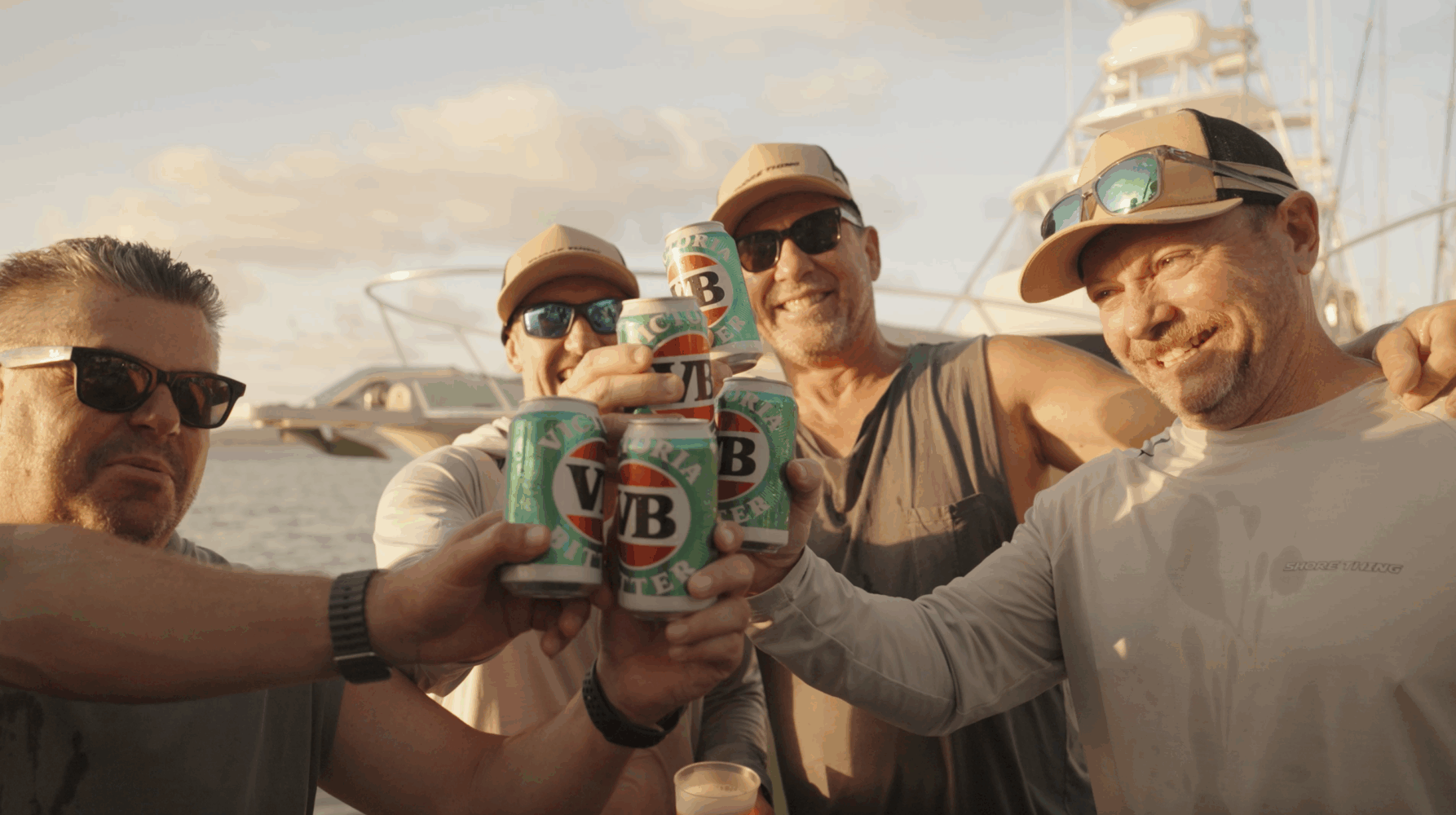Four men in sunglasses and casual clothing smile and toast with cans of Victoria Bitter beer on a boat, with sunny skies and another boat in the background.