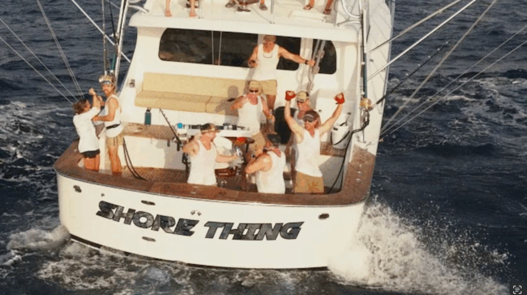 A group of people in white shirts celebrate on the deck of a boat named "Shore Thing" as it moves through the ocean, with water splashing at the stern.