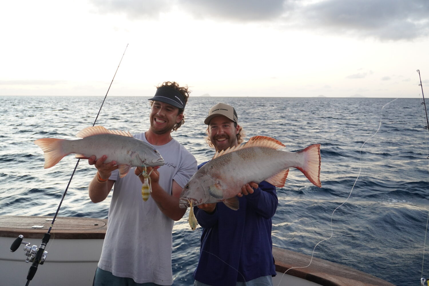Two smiling people on a boat hold large fish they caught, with the ocean and cloudy sky in the background. Fishing rods and deck rails are visible.