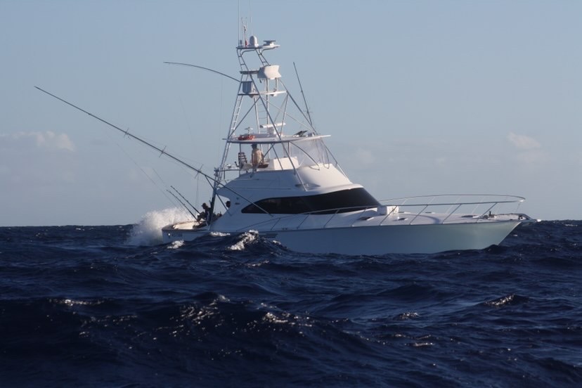 A white fishing yacht with tall antennas moves through choppy, dark blue ocean waves under a clear sky, with water splashing against its hull.