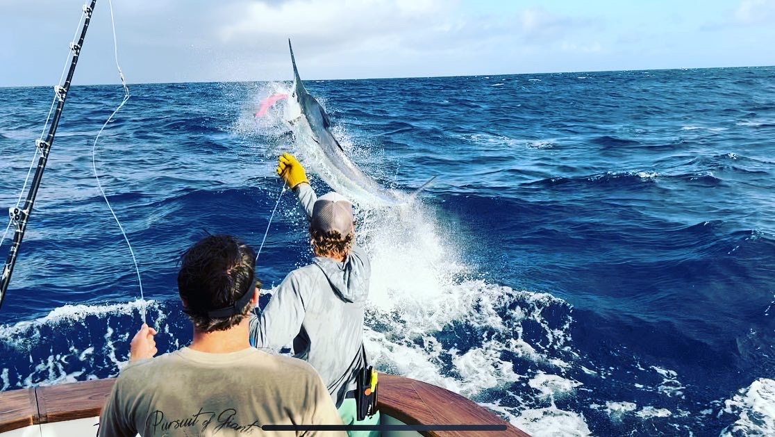 Two people on a boat are deep sea fishing as a large marlin leaps out of the ocean, creating a splash. One person is holding the fishing line while wearing gloves. The sky is partly cloudy.