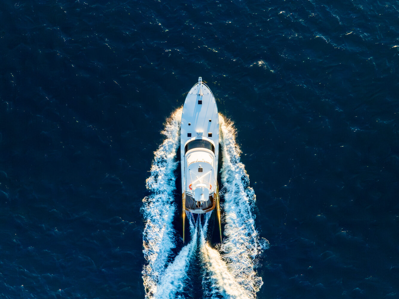 Aerial view of a white yacht cruising through deep blue water, leaving a foamy wake behind. The sunlight highlights the yacht's deck and the waves it creates on the calm sea.
