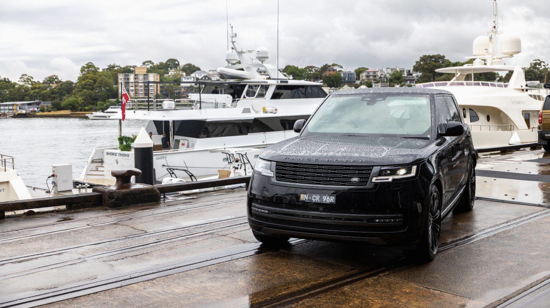 A black Range Rover SUV parked on a dockside near several luxury yachts, with water and buildings in the background on a cloudy day.