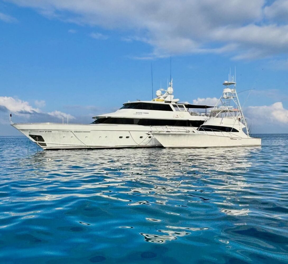 A large white luxury yacht floats on calm, blue water under a partly cloudy sky, with its reflection visible on the surface.