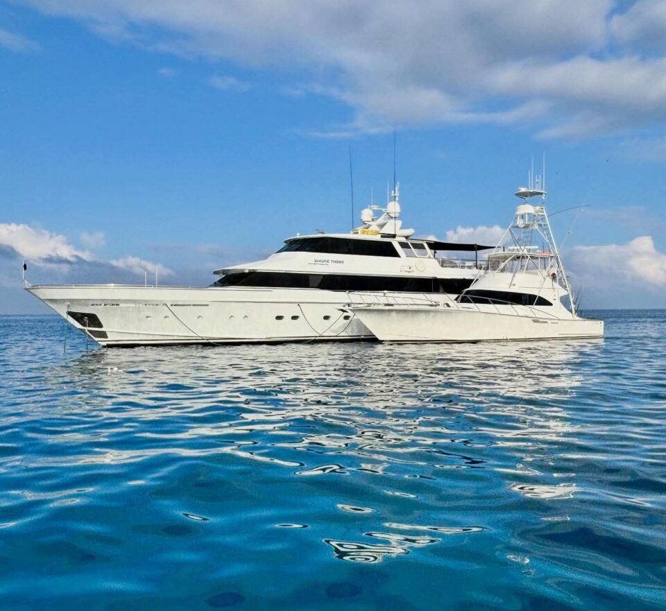 A large white luxury yacht floats on calm, blue water under a partly cloudy sky, with its reflection visible on the surface.