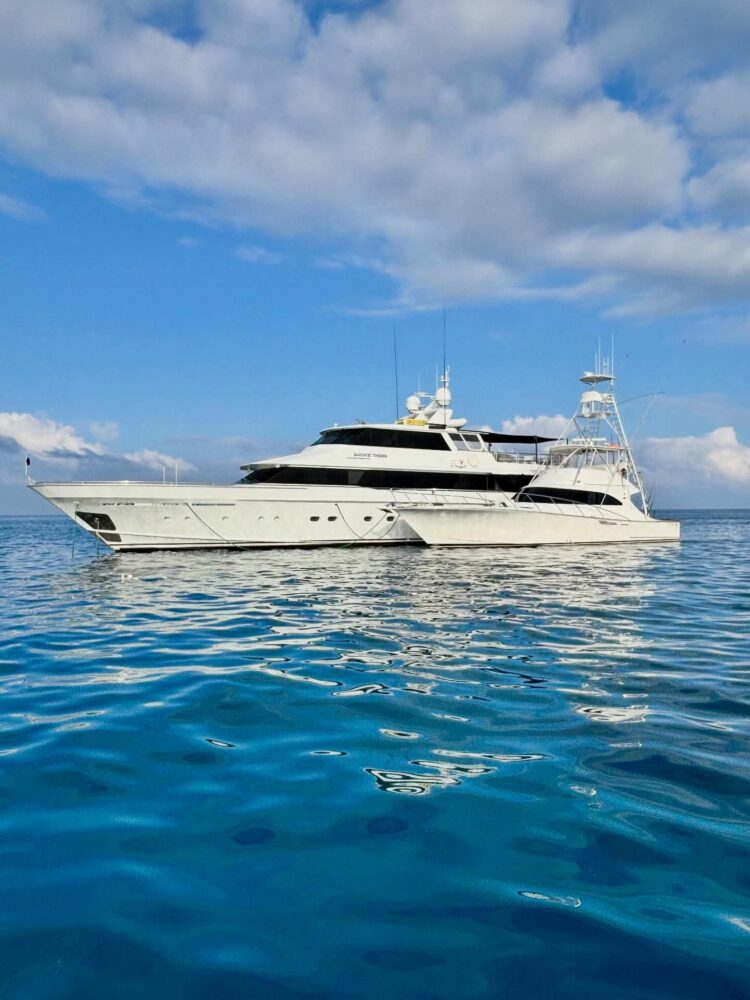 A large white luxury yacht floats on calm, clear blue water under a partly cloudy sky, with reflections of the boat and clouds visible on the sea's surface.