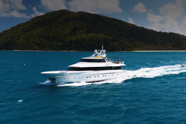 A large white yacht cruises through clear blue water near a lush, forested island under a partly cloudy sky.