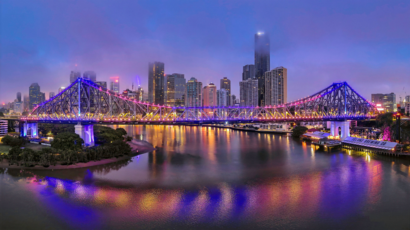 A city skyline at dusk with a brightly lit bridge spanning a river, colorful lights reflecting on the water, and tall buildings in the background under a cloudy sky.
