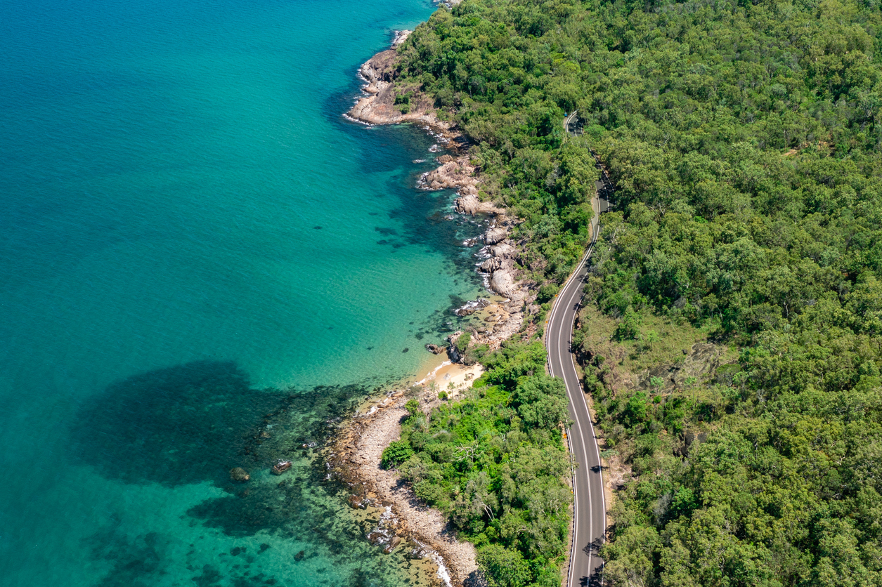A winding road runs along a lush green coastline with clear turquoise water, rocky shorelines, and dense forest beside the ocean.