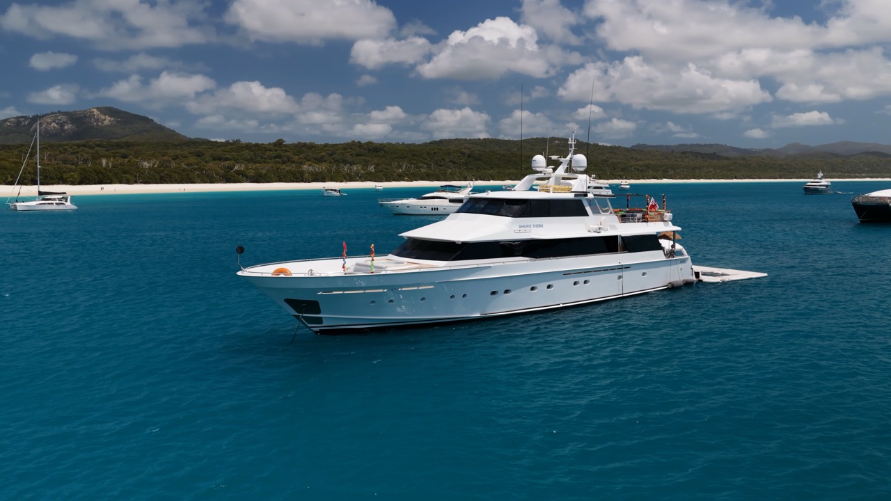 A white luxury yacht anchored in clear blue water near a sandy beach with green hills in the background and other boats nearby under a partly cloudy sky.
