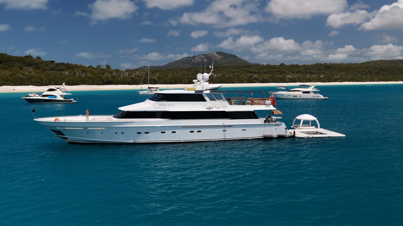 Luxury yacht anchored in clear blue water near a tropical island, with other boats and a sandy beach in the background under a partly cloudy sky.