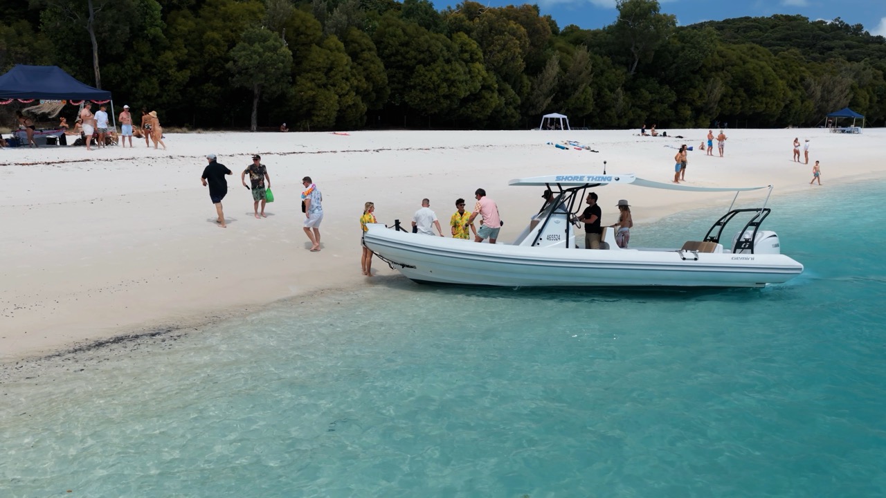 A group of people stands near a small white boat on a sandy beach with clear blue water. More people relax under tents and trees in the background. The scene is bright and tropical.