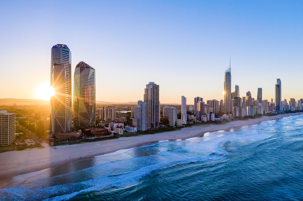 Aerial view of a city skyline with tall modern buildings along a sandy beach at sunrise, with gentle waves and clear blue water in the foreground.