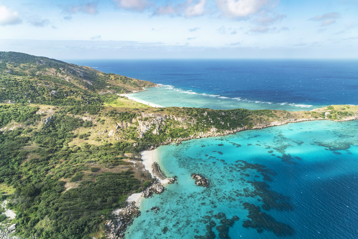 Aerial view of a tropical coastline with lush green hills, rocky shores, and clear turquoise ocean water under a partly cloudy sky.