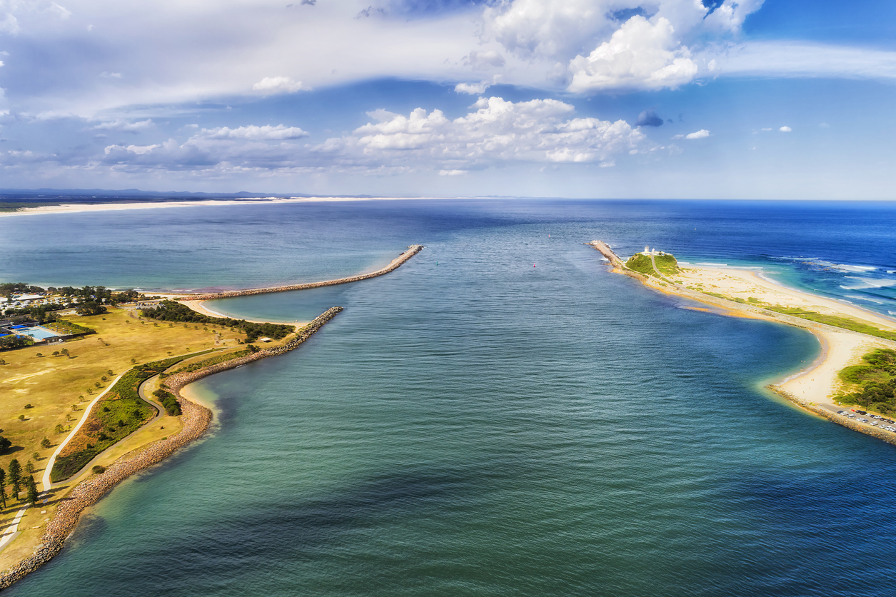 Aerial view of a river mouth meeting the ocean, with breakwaters on each side, grassy land, scattered trees, a lighthouse, and a partly cloudy sky above.