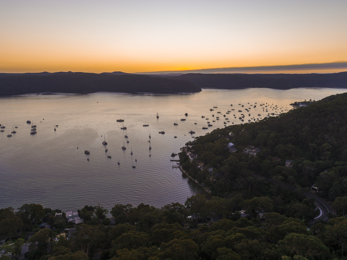 Aerial view of a calm bay at sunset with numerous boats anchored in the water, forested coastline, and distant hills under an orange sky.