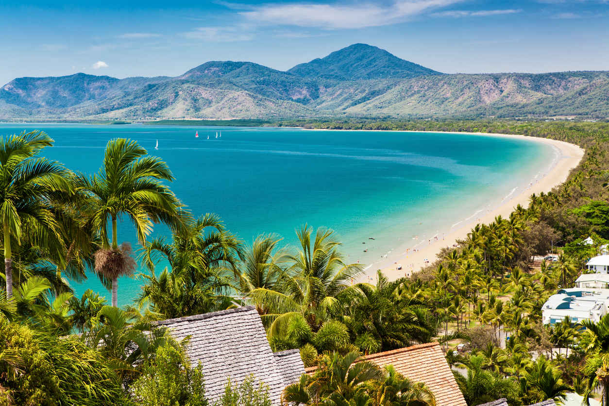 Scenic view of a tropical beach with turquoise water, white sand, palm trees, and distant green mountains under a blue sky. Sailboats are visible on the water.