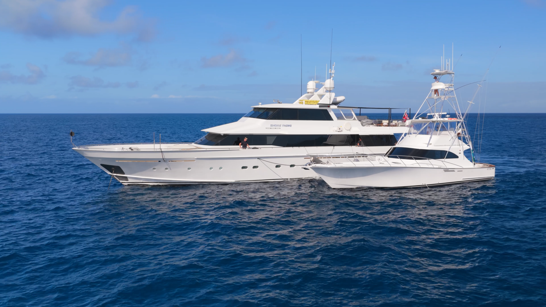 Two large white yachts are side by side on calm blue ocean water under a clear sky, with a few people visible on the decks.