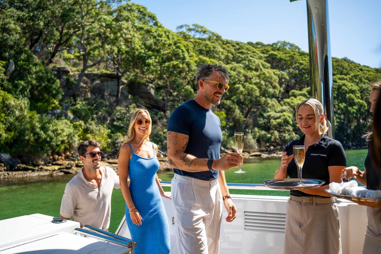 Four people smile on a boat by the water as a server hands out champagne. Trees and greenery are visible in the background under bright sunlight.