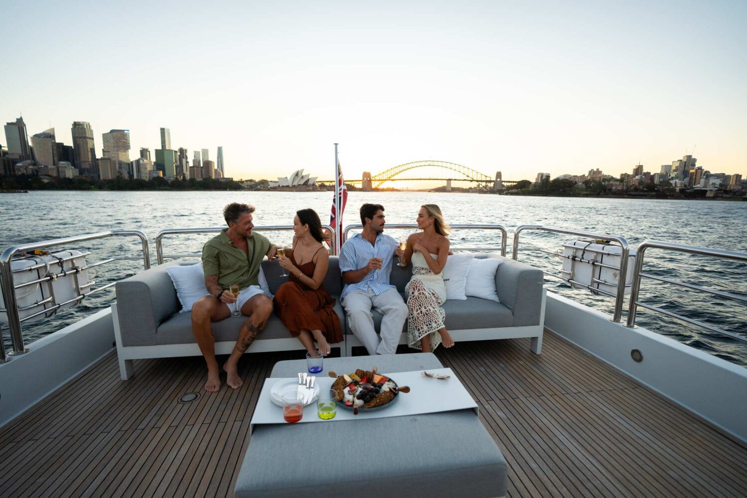 Four people sit on a couch on a yacht, enjoying drinks and snacks, with a city skyline and a bridge in the background at sunset.