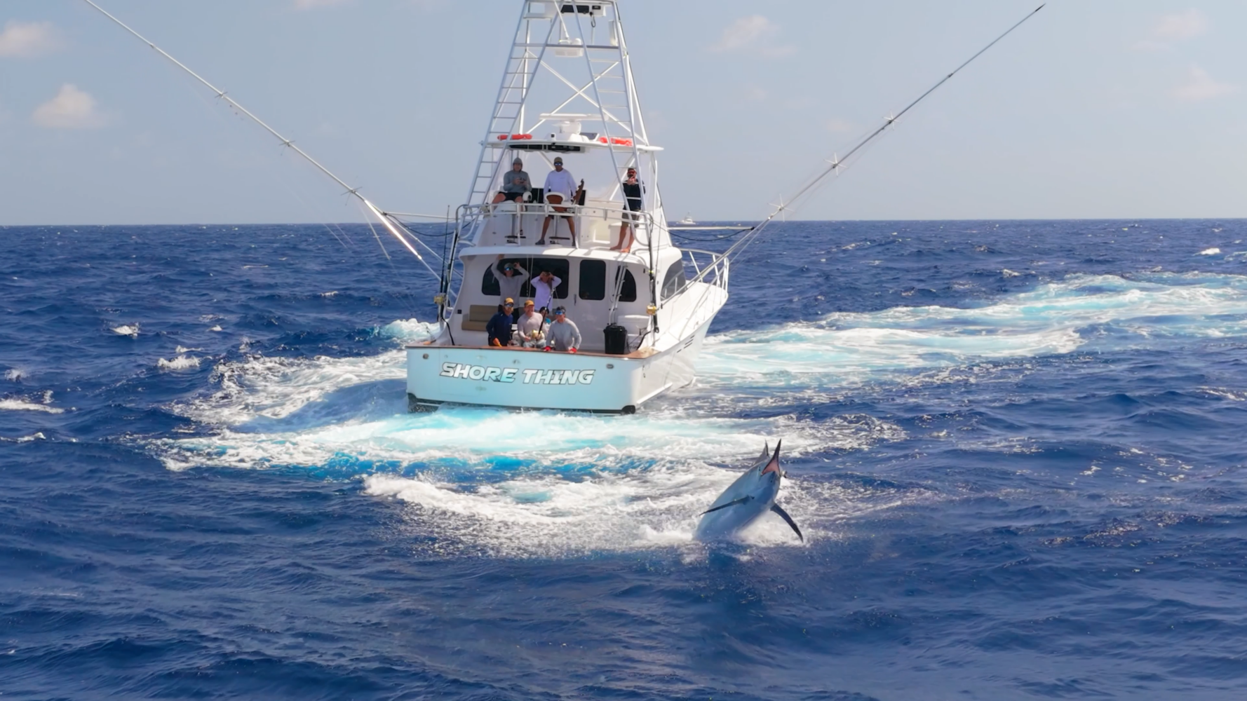 A fishing boat named “SHORE THING” on deep blue ocean water with several people aboard. A large fish leaps out of the water near the boat.