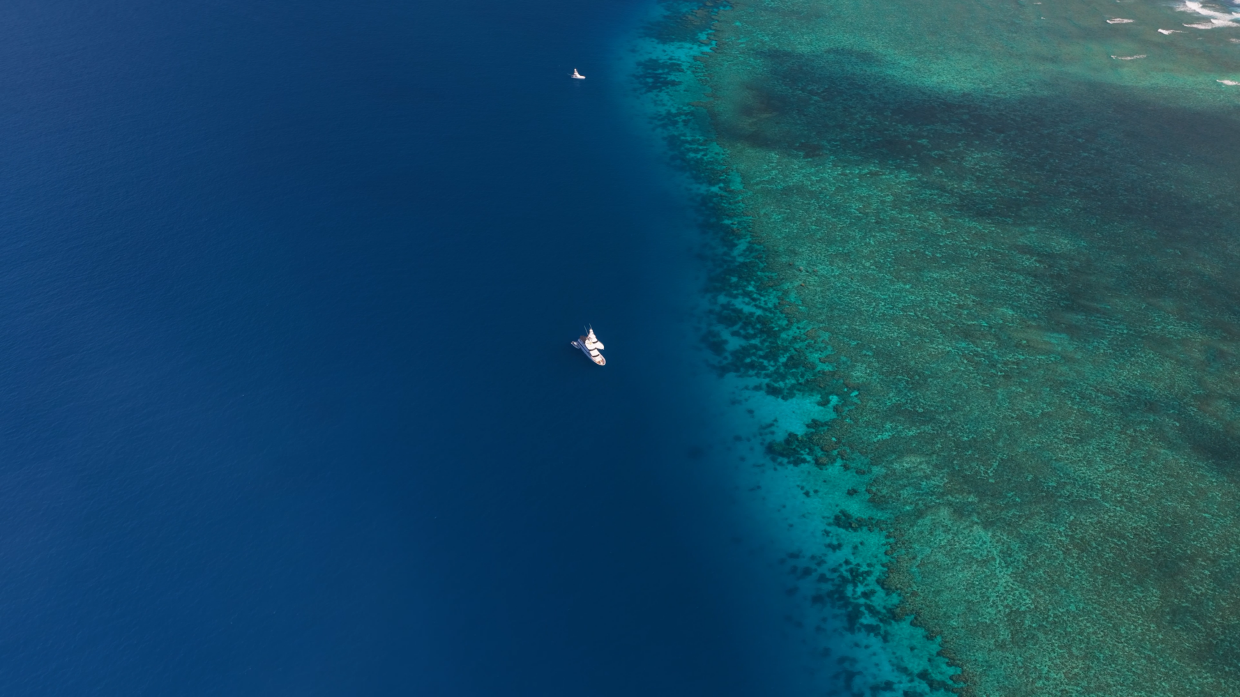 Aerial view of boats floating where deep blue ocean meets vibrant coral reef, highlighting the boundary between deeper and shallower water.