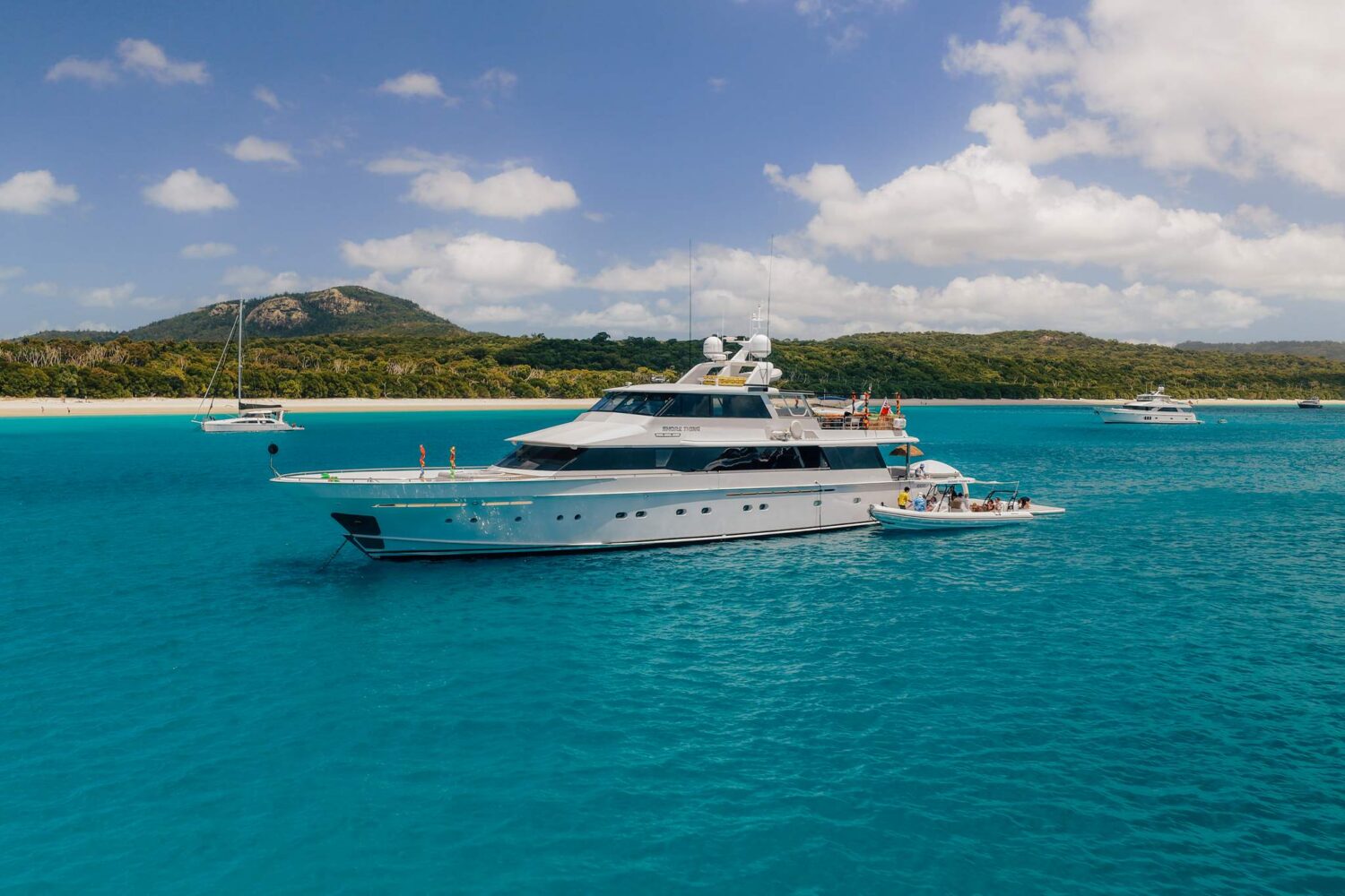 A large yacht anchored in clear blue water near a tropical island, with smaller boats nearby and people on board. Green hills and a sandy beach are visible in the background under a partly cloudy sky.