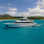 A large white yacht anchored in clear blue water near a sandy beach, with a small boat beside it and people on both vessels. Lush green hills and a partly cloudy sky are in the background.
