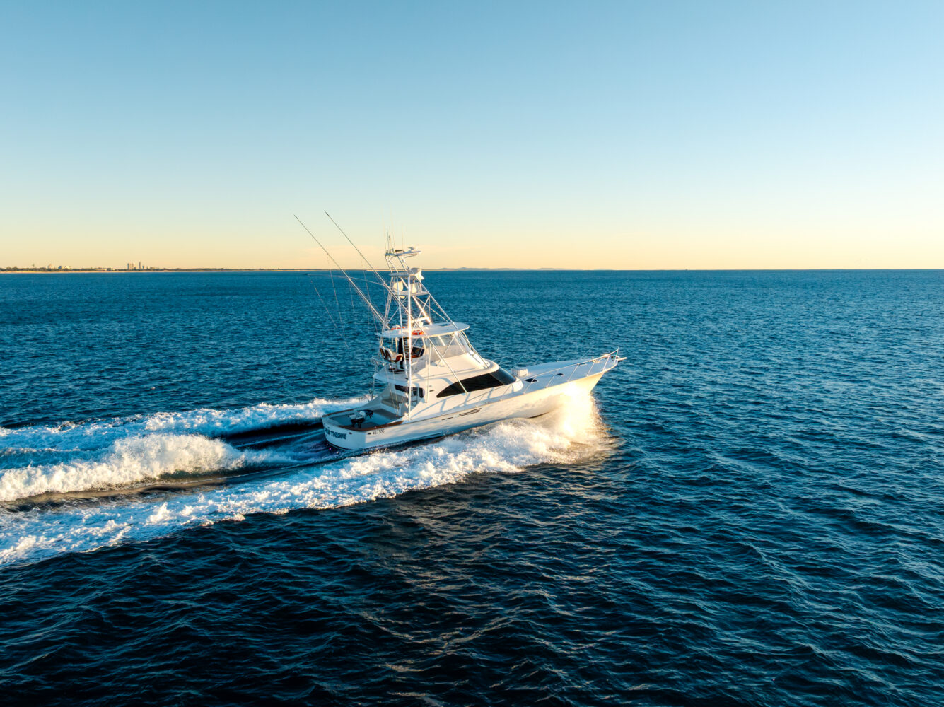 A white fishing boat cruises quickly across a calm, blue ocean under a clear sky at sunset, leaving a foamy wake behind.