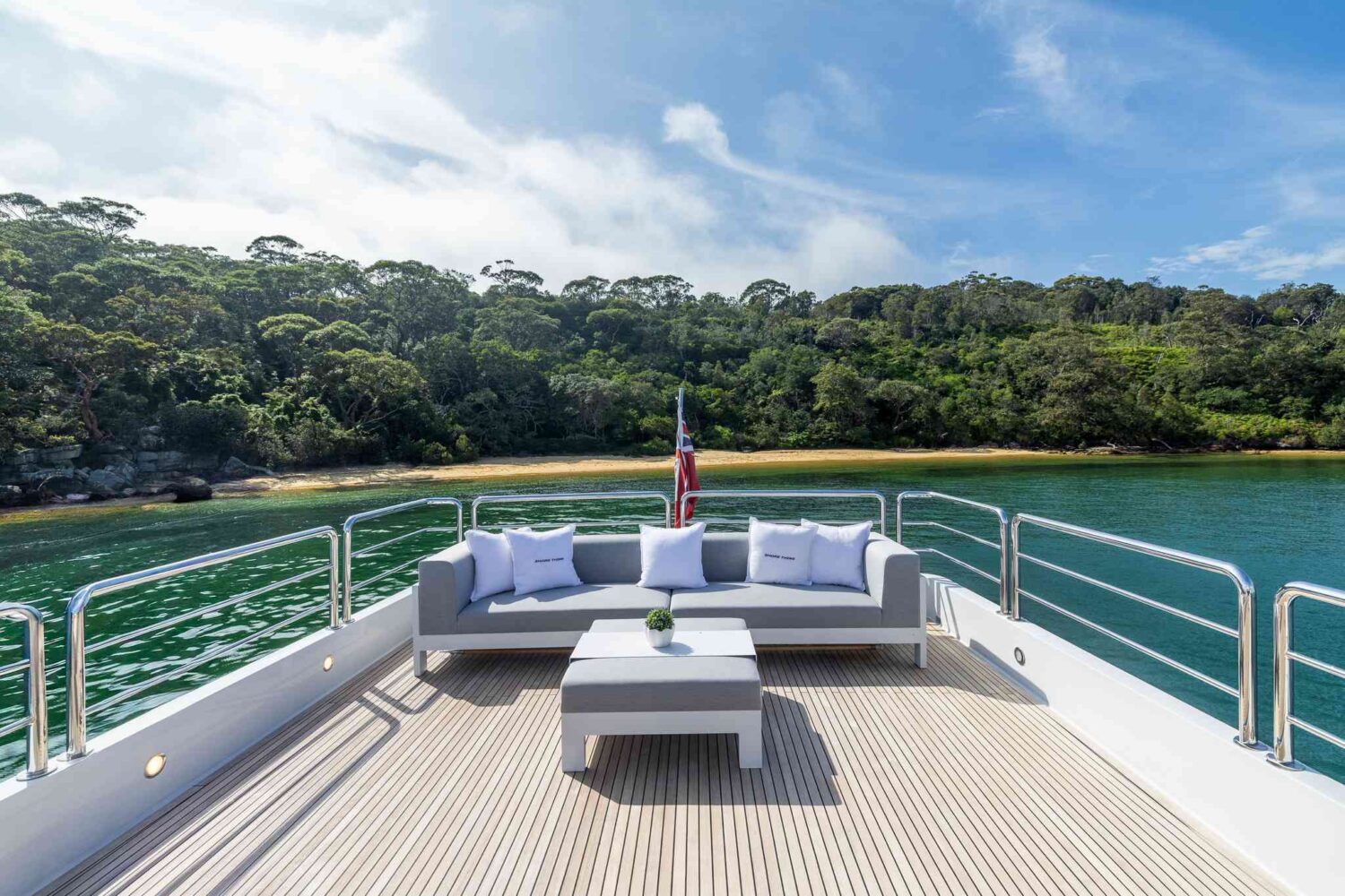 A yacht deck with a gray outdoor sofa, coffee table, and cushions faces a lush green shoreline and calm water under a partly cloudy sky. An American flag is visible at the back of the deck.