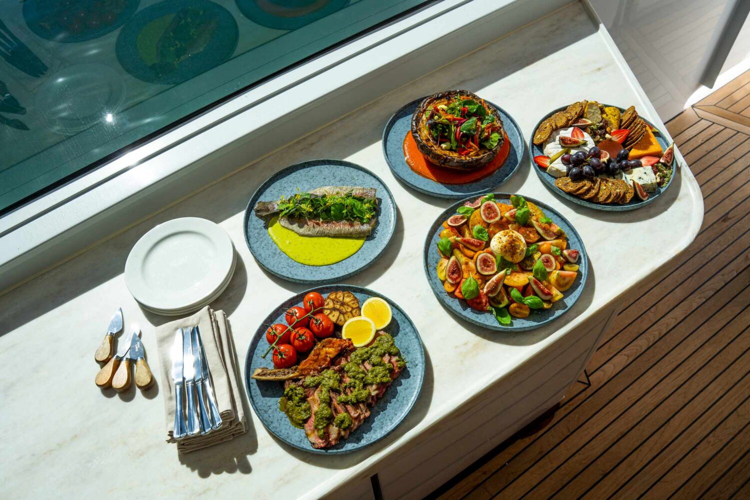 Six plates of colorful food, including grilled fish, steak with tomatoes, salad with figs, grilled vegetables, and fruit, are arranged on a white table with cutlery and plates beside them. Sunlight shines on the fresh dishes.