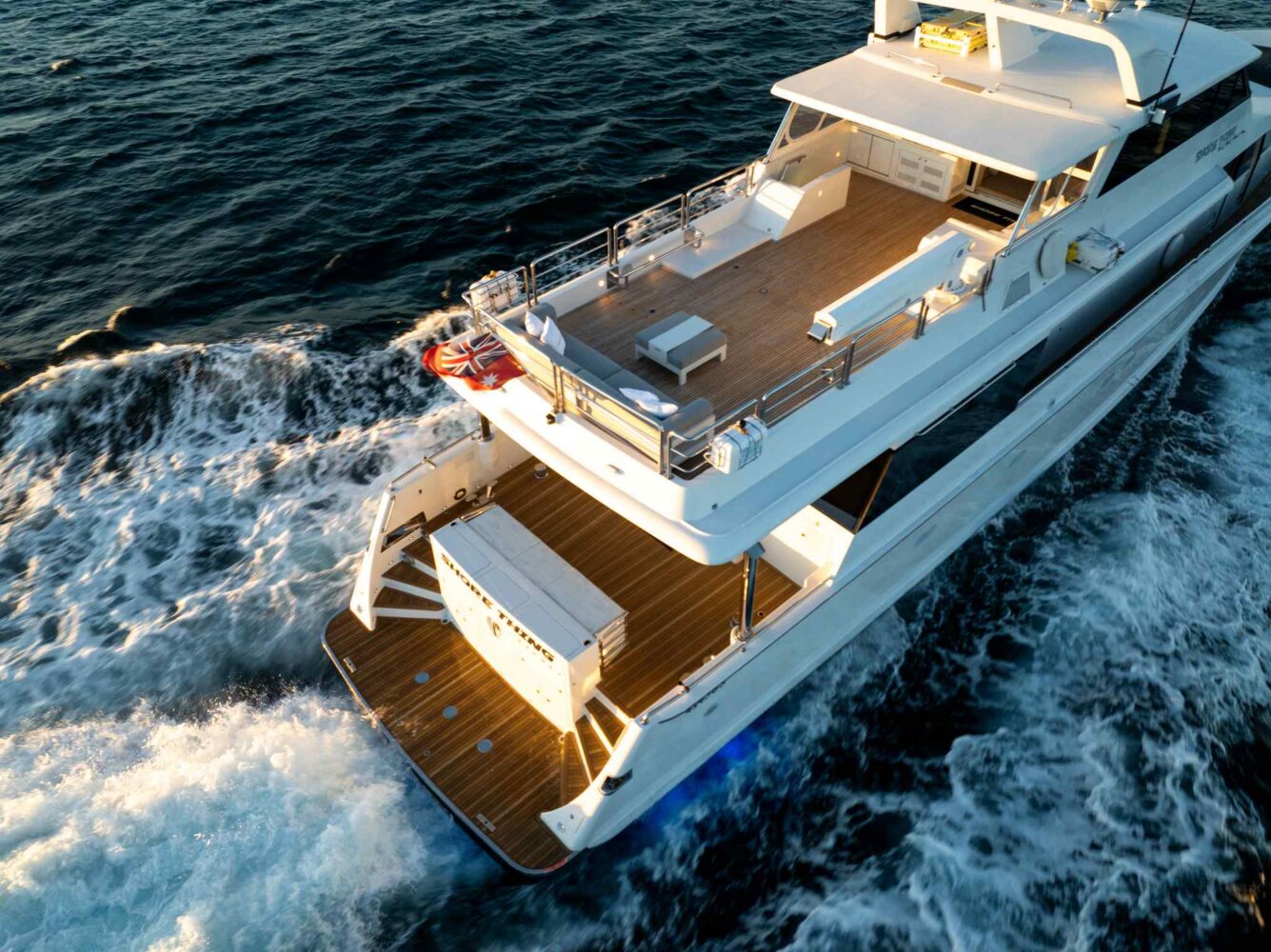 Aerial view of a luxury yacht cruising through the ocean, leaving a white wake behind. The yacht has a spacious wooden deck, lounge seating, and an open area at the back.
