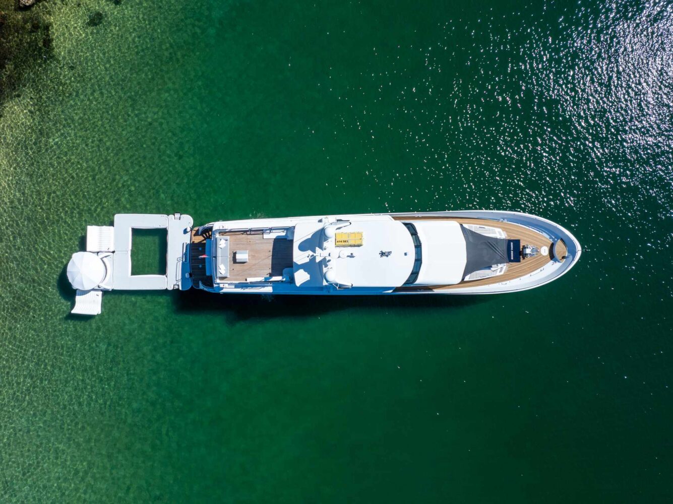 A large white yacht is docked in clear green water, viewed from above. A small lounge area with an umbrella is set up on a floating dock attached to the yacht’s stern.