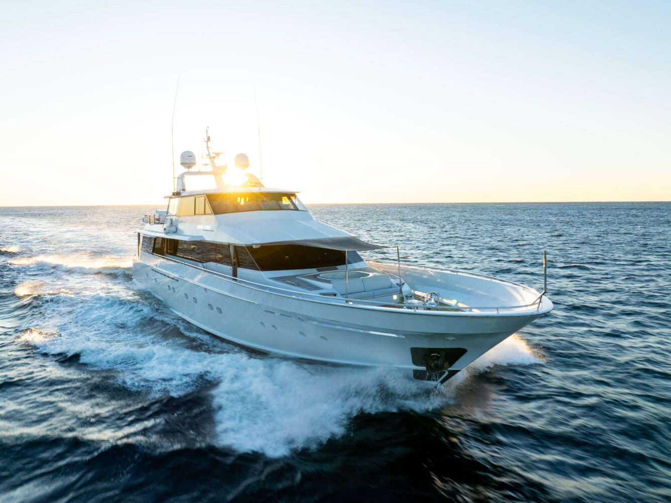 A large white yacht speeds across the ocean at sunset, creating waves as it moves forward, with the sun shining brightly in the background.