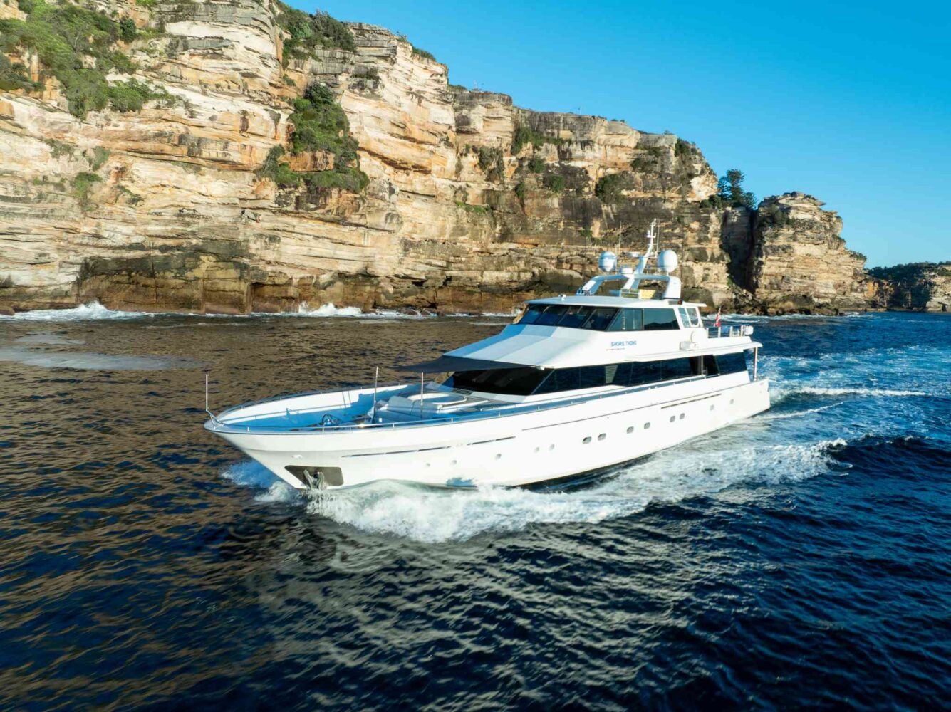 A white yacht cruises on blue water near rocky cliffs, with waves trailing behind it under a clear blue sky.