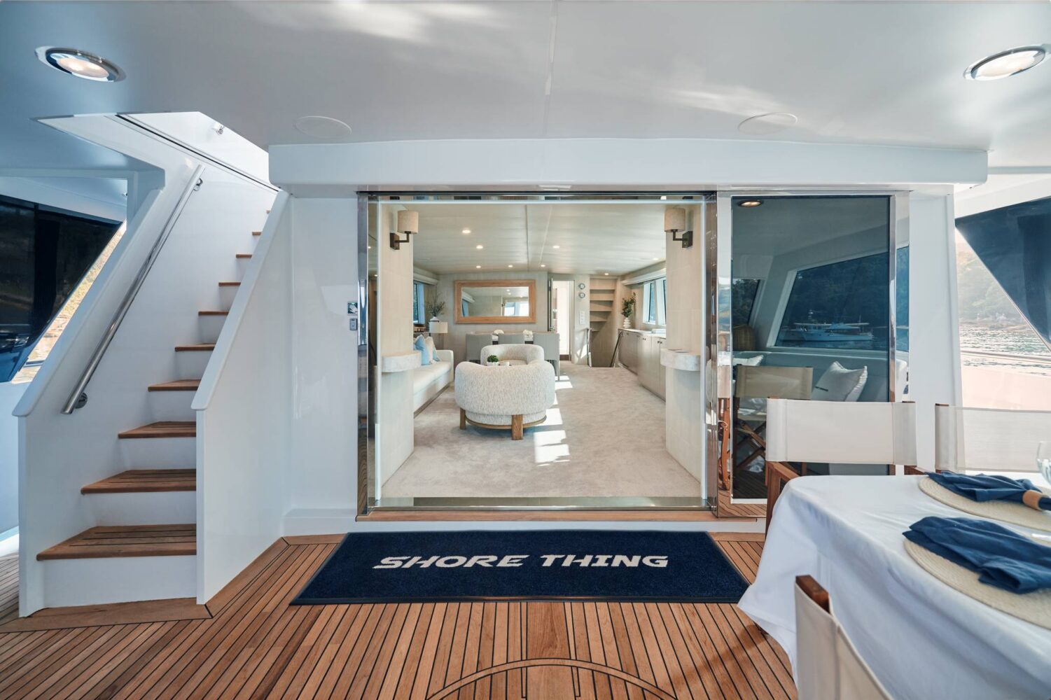 View of a yacht’s interior from the deck, with a doormat labeled “Shore Thing,” stairs to the left, and a dining table to the right. Large glass doors open to a modern, luxurious living space.