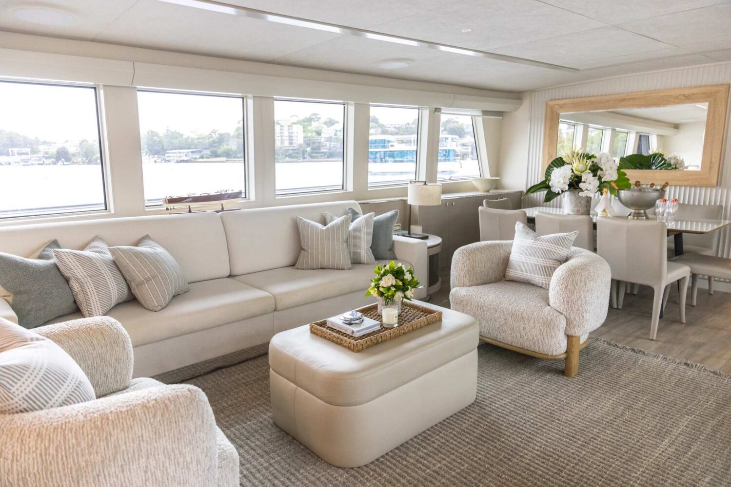 Modern yacht living area with cream sofas, cushioned chairs, a large ottoman, and a dining table. Large windows provide water views, and decorative plants and pillows enhance the neutral-toned, cozy space.