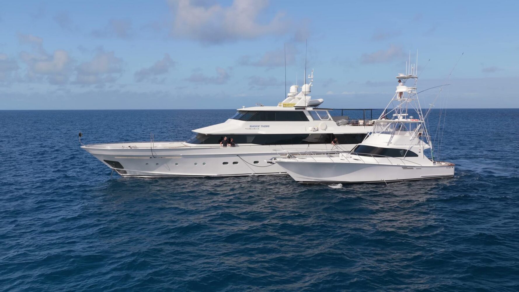 Two white yachts are anchored side by side on calm blue ocean water under a mostly clear sky with scattered clouds.