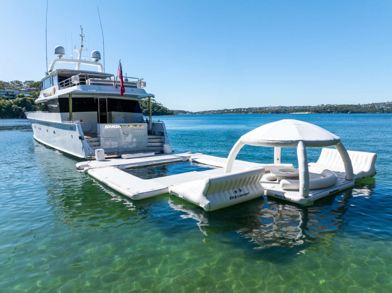 A large yacht is anchored in clear blue water next to a floating inflatable lounge with seats and a canopy. The shore and houses are visible in the background under a clear sky.
