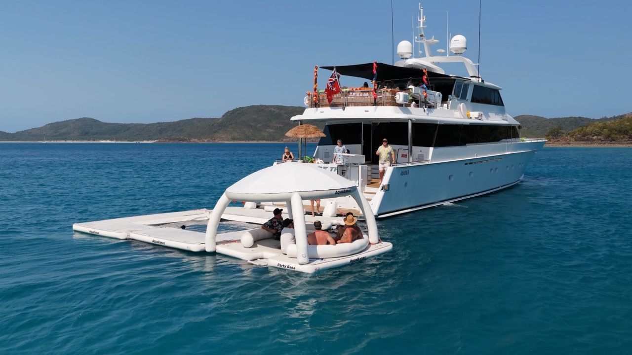 A large white yacht is anchored in clear blue water near green hills. Several people sit on a floating platform with a canopy beside the yacht, enjoying the sunny day.