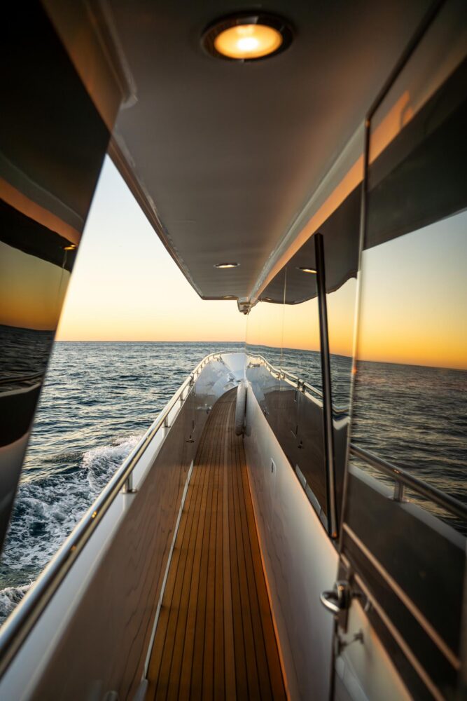 A view down the side deck of a yacht at sunset, with calm ocean waves and warm light reflecting off the yacht’s sleek surfaces.