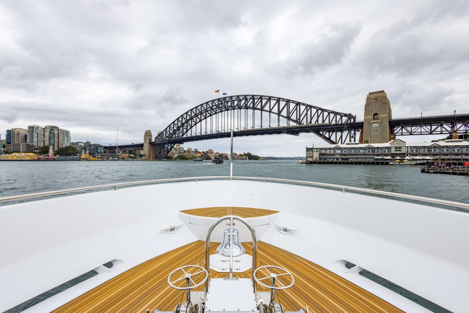 View from a yacht’s bow looking toward Sydney Harbour Bridge under a cloudy sky, with city buildings and waterfront structures visible on both sides of the harbor.